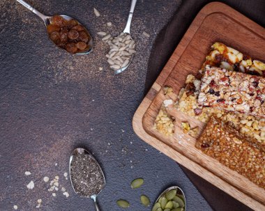 Kahverengi arka planda doğal granola çubukları ve kaşıklarda malzemeler.