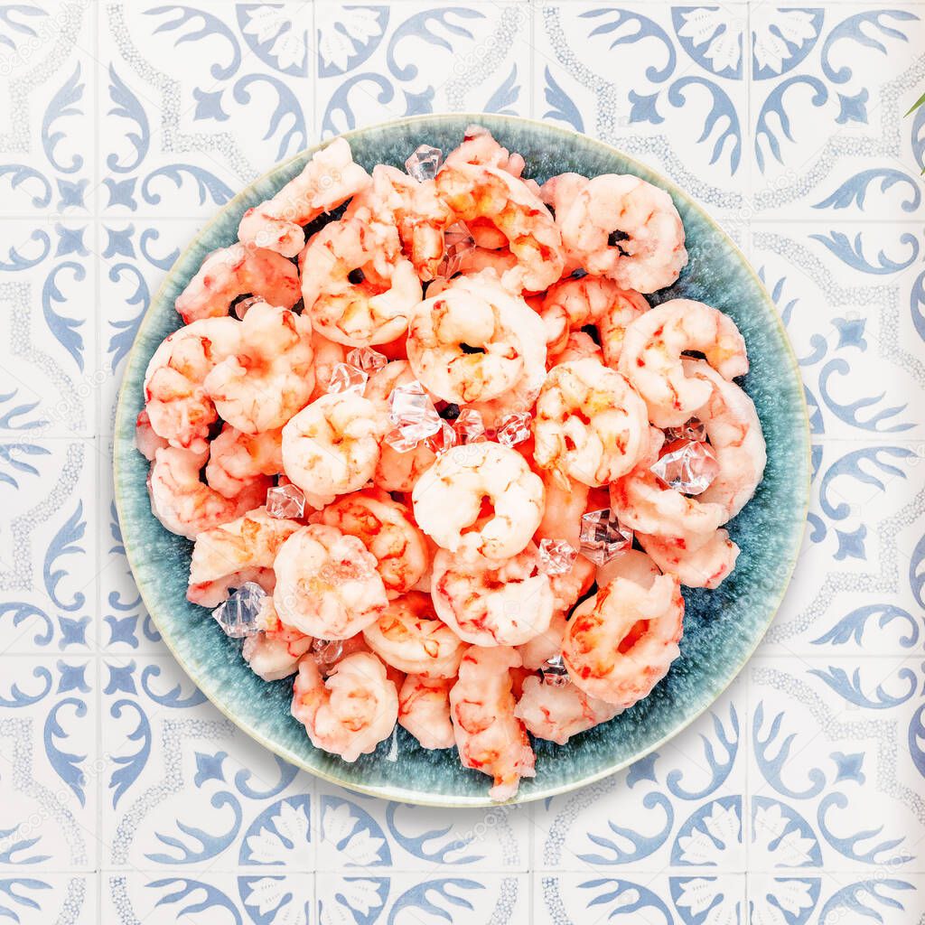 Close up natural frozen pilled shrimps with ice cubes on a blue plate on a tile ornamental table. Prawns cooking. Frozen foods. Mediterranean, keto diet