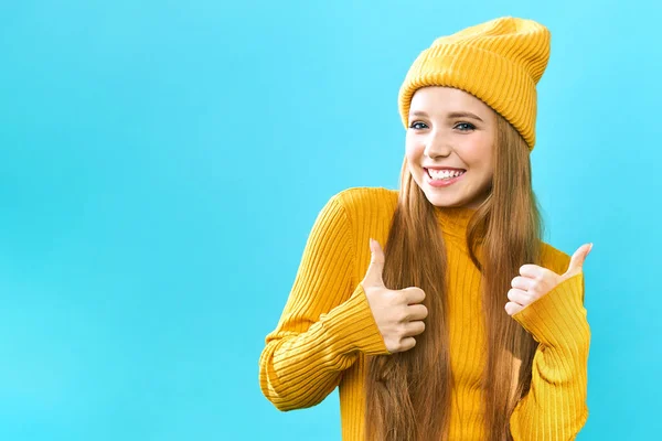 Portret van een jonge vrouw die blij is haar duim omhoog te laten zien. Glimlachend meisje met een gele hoed en gele trui op een blauwe achtergrond. Winkelen tijdens een grote Black Friday sale. — Stockfoto