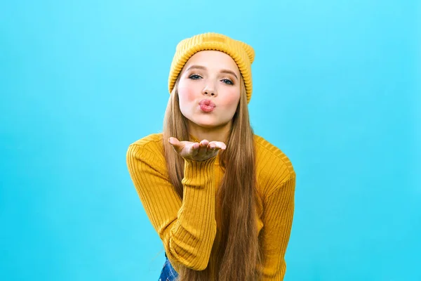 Portrét mladé ženy, která se raduje a posílá polibek. Usmívající se dívka ve žlutém klobouku a žlutém svetru na modrém pozadí. Nakupování během velkého prodeje Black Friday. — Stock fotografie