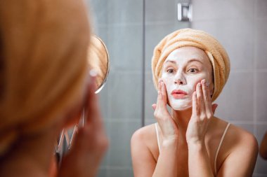 Bir kadın banyoda aynanın önünde yüzüne temizleyici bir maske takıyor. Cilt bakımı. Kozmetik kaplıca tedavileri.