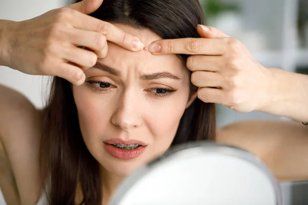 Una mujer aplasta una espinilla mientras se mira en el espejo. Imágenes de stock libres de derechos