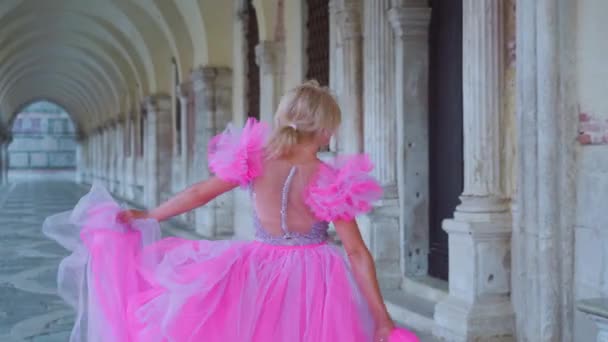 Blondýny dívka v elegantních růžových šatech chodí zezadu mezi benátskými sloupy