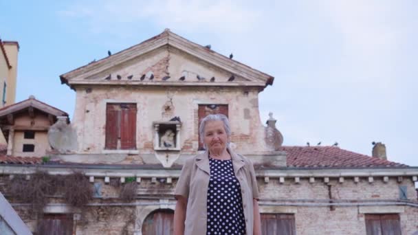 Пожилая женщина стоит напротив старого заброшенного здания — стоковое видео