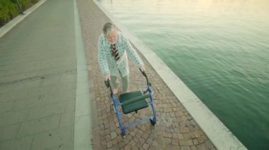Gri saçlı yaşlı kadın lagünün yanında rollator kullanarak yürüyor.