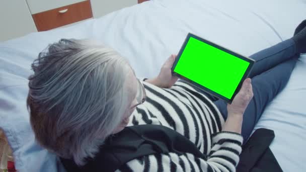 Staruszka czyta książki elektroniczne za pomocą tabletu leżącego na łóżku — Wideo stockowe