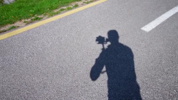 Χειριστής άνθρωπος κινείται κάμερα γυρίσματα ασφαλτοστρωμένο δρόμο κοντά λιβάδι — Αρχείο Βίντεο