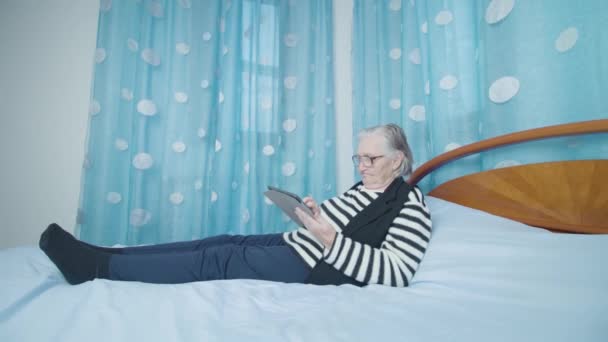 Vieja mujer de pelo gris lee noticias a través de una tableta acostada en la cama — Vídeos de Stock