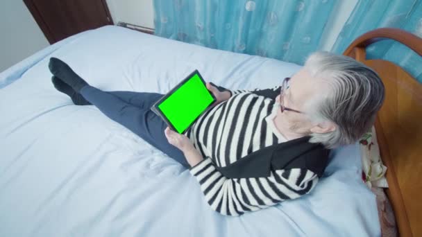 Egy ősz hajú nő híreket olvas tablettával az ágyon fekve. — Stock videók