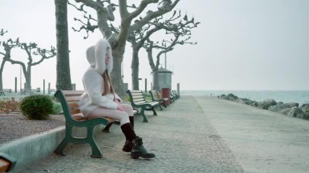 毛皮で覆われたフードを着た白いコートの女性は ガルダ湖の堤防の裸の木に対して木製のベンチに座っています 観光都市ペスキエーラ ガルダの曇りの天気 — ストック動画