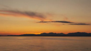 Garda Gölü, gün batımında karanlık dağ siluetine karşı. Dumanlı bulutlu turuncu gökyüzü alacakaranlık saatinde berrak suya yansıyor