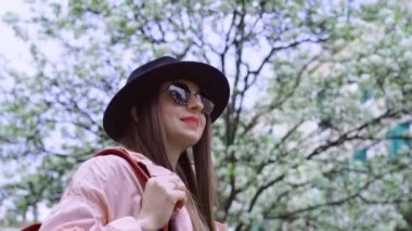 Siyah şapkalı ve güneş gözlüklü bir kadın ağaçların önünde dikilip etrafına bakıyor. Zarif bayan, İtalya 'da bahar tatilinde doğanın tadını çıkarıyor.