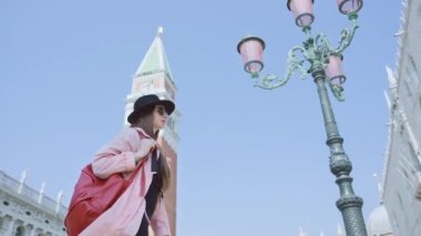 Şık giyinmiş bir kadın, St. Mark Meydanı 'nda mavi gökyüzünün altında sokak fenerinin önünden geçiyor. Bayan klasik İtalyan mimarisini inceliyor.