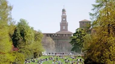 MILAN , ITALY - APRIL 06 2022: Tourists rest on grass against ancient building in Parch Sempion with lush green trees. Historical center of city on sunny day on April 06 in Milan
