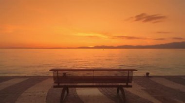 Wooden bench installed on city waterfront against Lake Garda at bright sunset. Orange light illuminates clear sky reflecting on water surface
