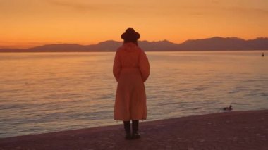 Zarif bayan İtalya 'daki Garda Gölü' nde inanılmaz günbatımının tadını çıkarıyor. Kadın şehrin rıhtımında durmuş su yüzeyine yansıyan aydınlık gökyüzüne bakıyor.