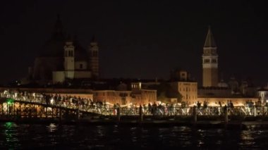 People walk on long pontoon bridge against illuminated historical buildings and street lights. Venetians celebrate Feast of Redeemer in evening