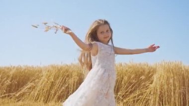 Cheerful girl has fun in ripe wheat field holding bouquet from golden spikelets. Happy child with long loose hair dances against clear blue sky