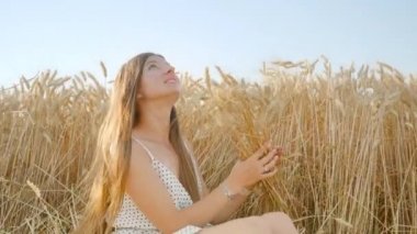 Smiling woman with long hair makes bouquet from wheat spikelets sitting in wheat field. Young female enjoys nature in countryside closeup