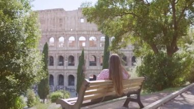 Woman looks at large Colosseum relaxing on bench under trees lacy shadow. Female traveler enjoys ancient architecture of Rome backside view