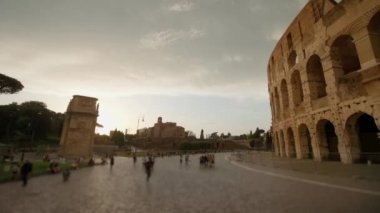 People walk near ancient Colosseum at sunset in Rome. Large amphitheater built in center of Italian capital attracts tourists on cloudy day