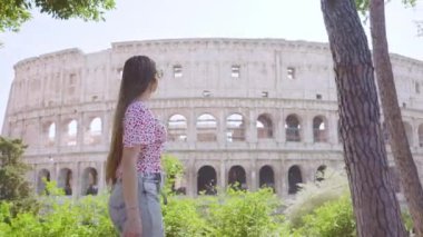 Young woman walks against large Colosseum behind green trees and bushes. Female tourist explores attractions of Rome on sunny summer day