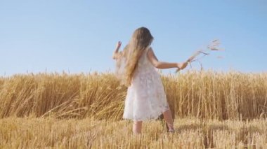 Cheerful girl dances in golden wheat field holding bouquet of spikelets. Cute child with long loose hair has fun in countryside on sunny day