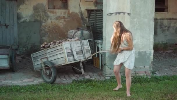 若い女性は荒廃した建物に対して庭の薪と古い木製のカートを引っ張る 長髪の疲れた女性は村で週末を過ごします — ストック動画