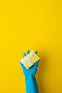 Mavi koruyucu lastik eldivenli bir el sarı arka planda köpüklü bir çamaşır süngeri tutar.