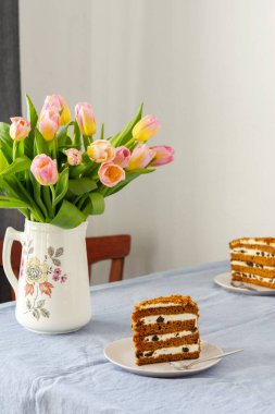 Bir sürahide pembe lale buketi ve masasında mavi bir masa örtüsü olan bir tabakta kuru erikli pasta.
