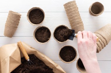 Tohumlar için çevre dostu tencereler ve beyaz ahşap arka planda zemin olan bir çanta, bahçe küreği olan kadın elleri, saksılara tohum ekmek için toprak döken bahçıvan, üst manzara.