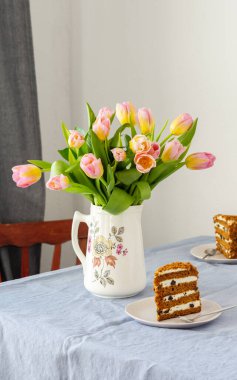 Bir sürahide pembe lale buketi ve masasında mavi bir masa örtüsü olan bir tabakta kuru erikli pasta.