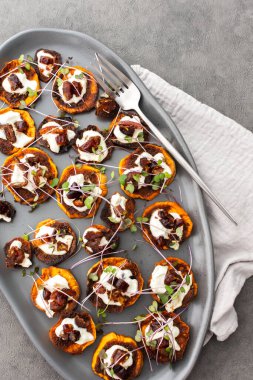 Fırında fırınlanmış portakallı tatlı patates yoğurt ve hurma, mikro yeşillikler serpiştirilmiş, gri oval seramik tabakta tatlı patates, üst manzara.