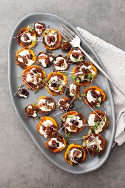 Fırında fırınlanmış portakallı tatlı patates yoğurt ve hurma, mikro yeşillikler serpiştirilmiş, gri oval seramik tabakta tatlı patates, üst manzara.