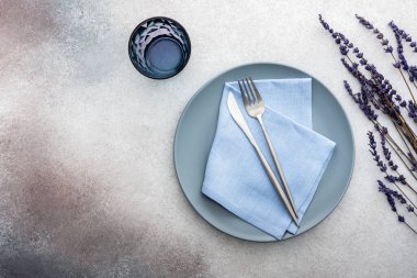 Masa ayarı, gri-pembe beton zemin üzerinde peçete ve çatal bıçaklı boş tabak, servis edilen masanın üst manzarası kuru lavanta çiçekleriyle süslenmiş.