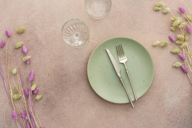 Masa ayarı, boş yeşil tabak ve beton zemin üzerinde çatal bıçak takımı, servis edilen tablonun üst görüntüsü kuru çiçeklerle süslenmiş.