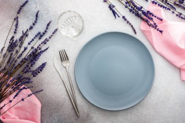 Masa ayarı, gri-pembe beton zemin üzerinde peçete ve çatal bıçaklı boş tabak, servis edilen masanın üst manzarası kuru lavanta çiçekleriyle süslenmiş.