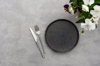 Masa ayarı, boş tabak ve gri arka planda çatal bıçak takımı, servis edilen masanın üst görüntüsü hanım evladı çiçekleriyle süslenmiş.