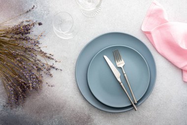 Masa ayarı, gri-pembe beton zemin üzerinde peçete ve çatal bıçaklı boş tabak, servis edilen masanın üst manzarası kuru lavanta çiçekleriyle süslenmiş.
