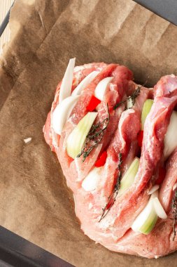 A large piece of raw pork meat prepared for baking, with onions and spices and thyme on parchment paper on a baking sheet