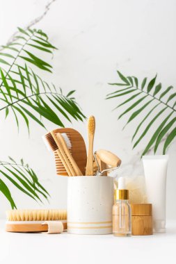 Seramik bardakta doğal tüyleri olan tahta diş fırçaları, yüz ve cilt bakım ürünleri, pipetli şişeler ve bir kavanoz krem, banyo aksesuarları, spa ve güzellik konsepti.