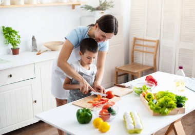 Bonding Concept. Cheerful mother helping her son to cook, teaching little boy how to prepare healthy vegetable salad, cutting tomatoes, cooking together at home.