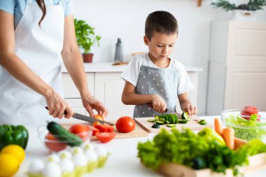 Oğlu evde annesine yardım ediyor. Çocuk annesiyle birlikte beyaz modern mutfakta yemek pişiriyor, salata için salatalık kesiyor. Birliktelik konsepti. Sağlıklı yemek..