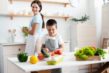 Cheerful mother proud her self-dependent son. Child unaided is cooking salad at kittchen. Healthy food concept.