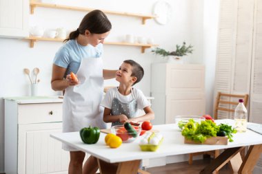 Cheerful mother proud her self-dependent son. Child unaided is cooking salad at kittchen. Healthy food concept.