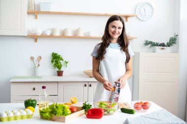 Gülümseyen kadın aşçı beyaz önlük giyen aşçı ev mutfağında sağlıklı salata pişiren sebzelerle masanın yanında duruyor.