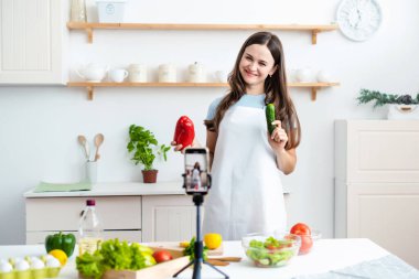 Food, culinary bloggers concept. Smiling young woman food blogger wearing apron and recording her cooking by mobile phone on tripod in kitchen, gesturing at phone camera, healthy fresh salad