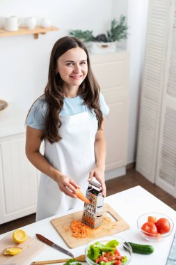 Masanın yanında sebzelerle duran beyaz önlüklü gülümseyen kadın ev mutfağında sağlıklı salata pişirmeye hazır.