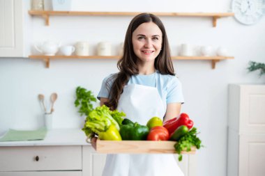 Beyaz önlük giyen gülümseyen aşçı kadın elinde sağlıklı taze sebzelerle ahşap tepsi tutuyor. Yemek, aşçılık blogcuları konsepti.