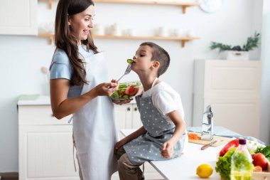 Mutlu gülümseyen ebeveynler küçük çocukla hafta sonu oyunlarının keyfini çıkarın. Anne ve oğul önlük giyip mutfakta yemek pişiriyor ve yemek tadıyorlar..